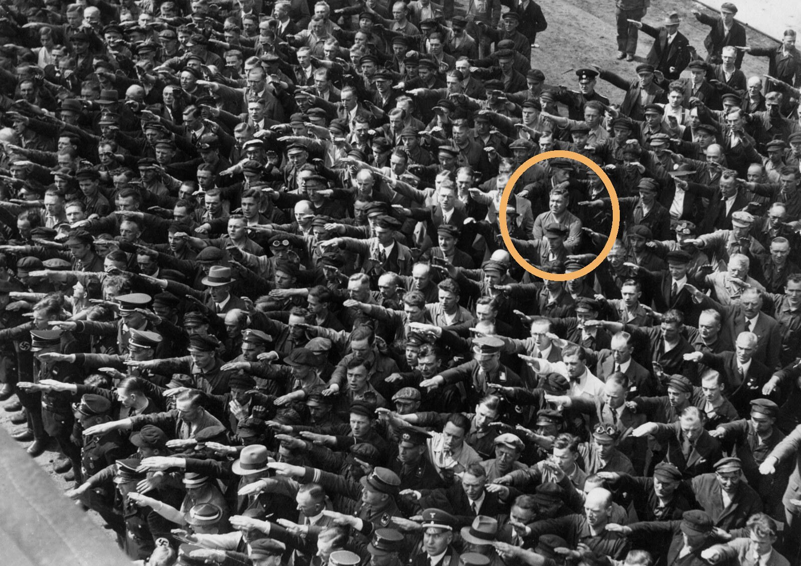 ภาพขาวดำปี 1936 ฝูงชนที่อู่ต่อเรือในฮัมบูร์กกำลังยกมือทำความเคารพแบบนาซี มีชายคนหนึ่งในวงกลม (เชื่อกันว่าคือ August Landmesser) ยืนกอดอกไม่ยอมยกมือ